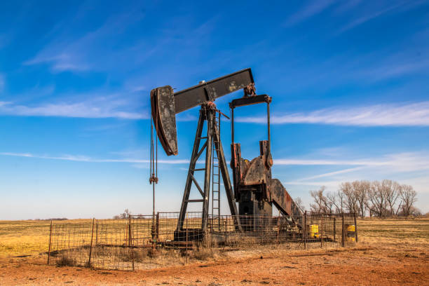 Large old rusty oil well pump jack surrounded by cattle panel fence out in field with very blue sky in winter.